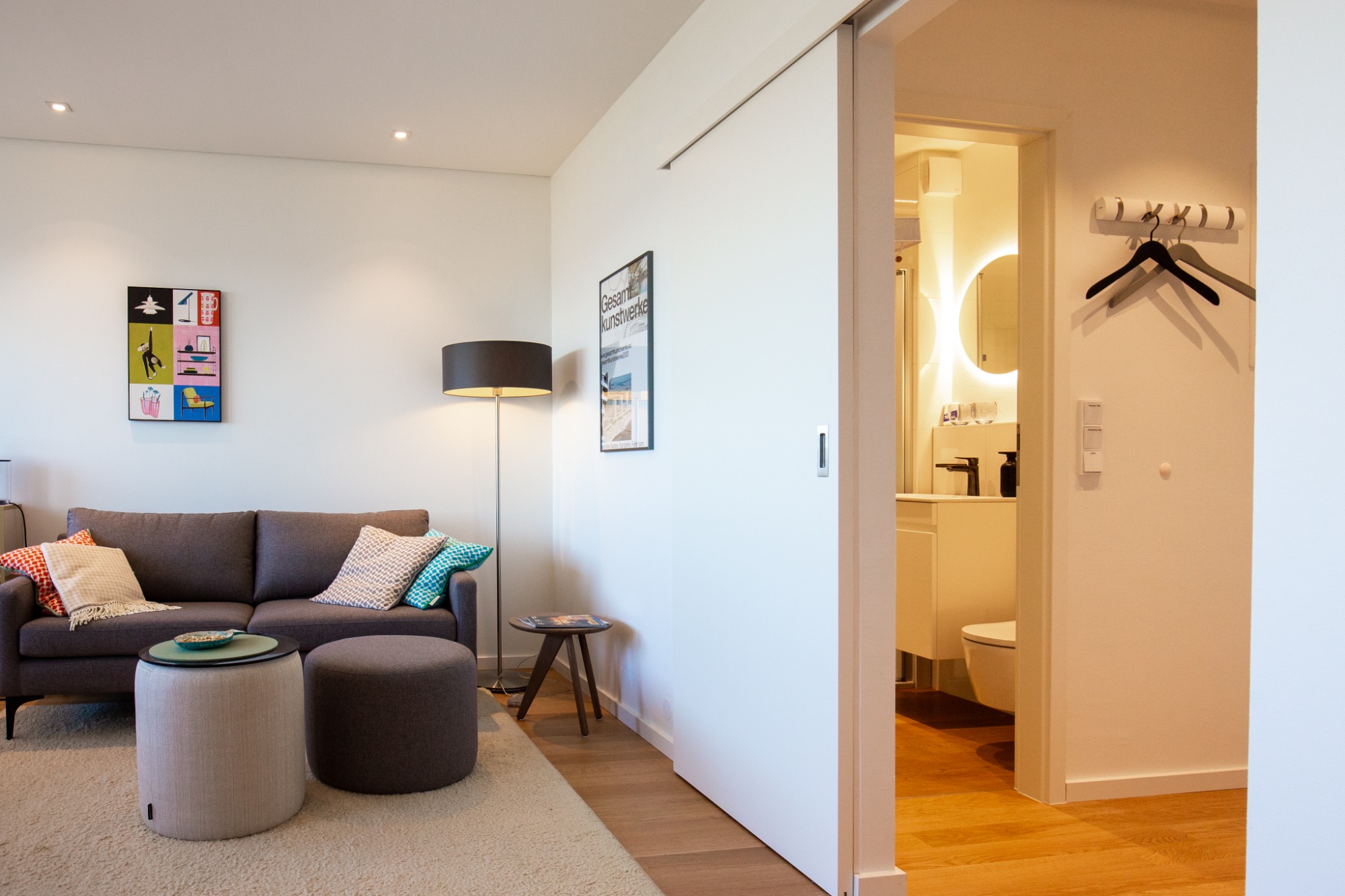 Entspannt-modernes Apartment Wohnzimmer mit grauem Sofa, Stehlampe, Hockern und Zugang zu einem Badezimmer.