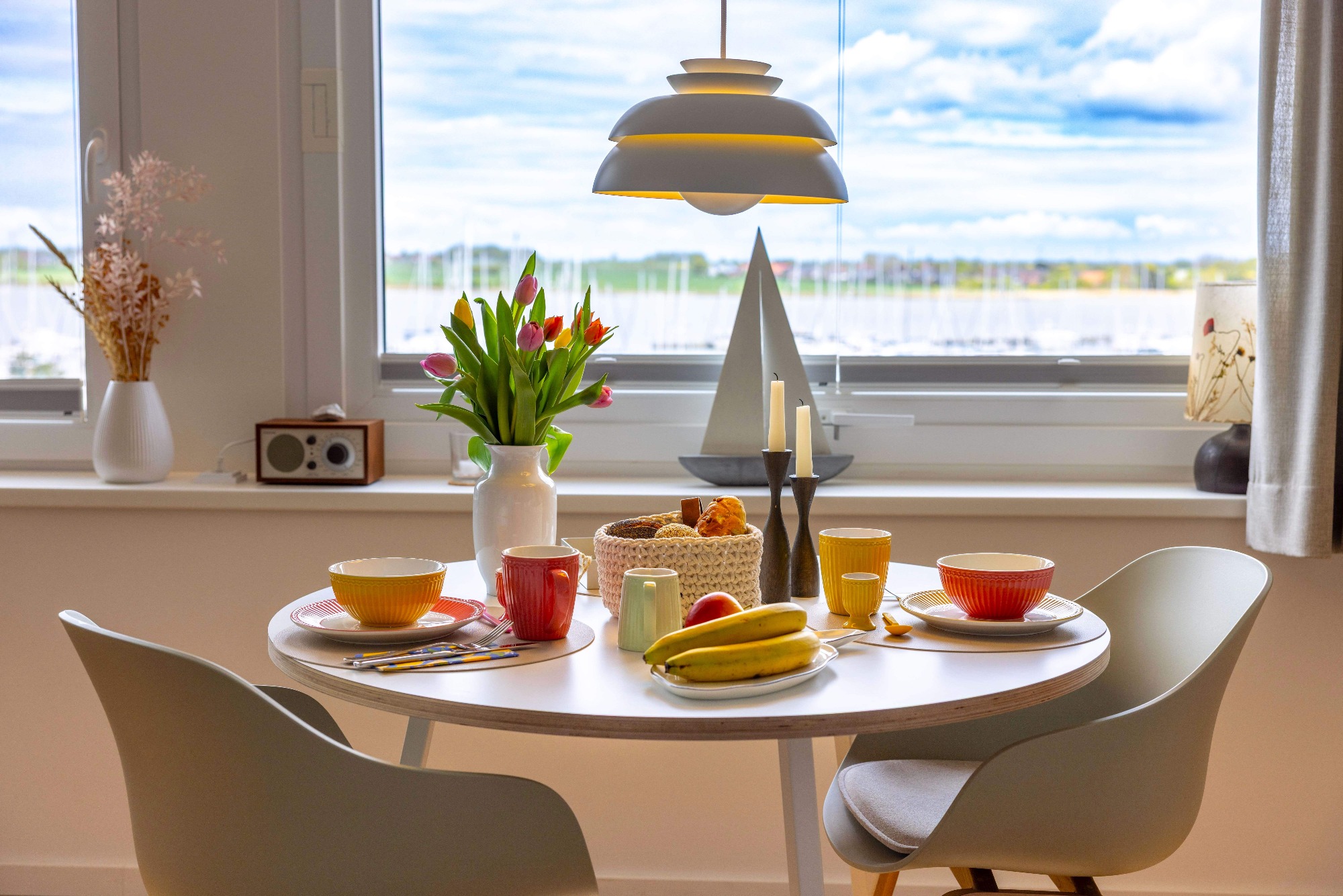 Sonne zum Frühstück Ein gedeckter Tisch mit frischen Blumen, Obst und Geschirr, vor einem Fenster mit Aussicht.