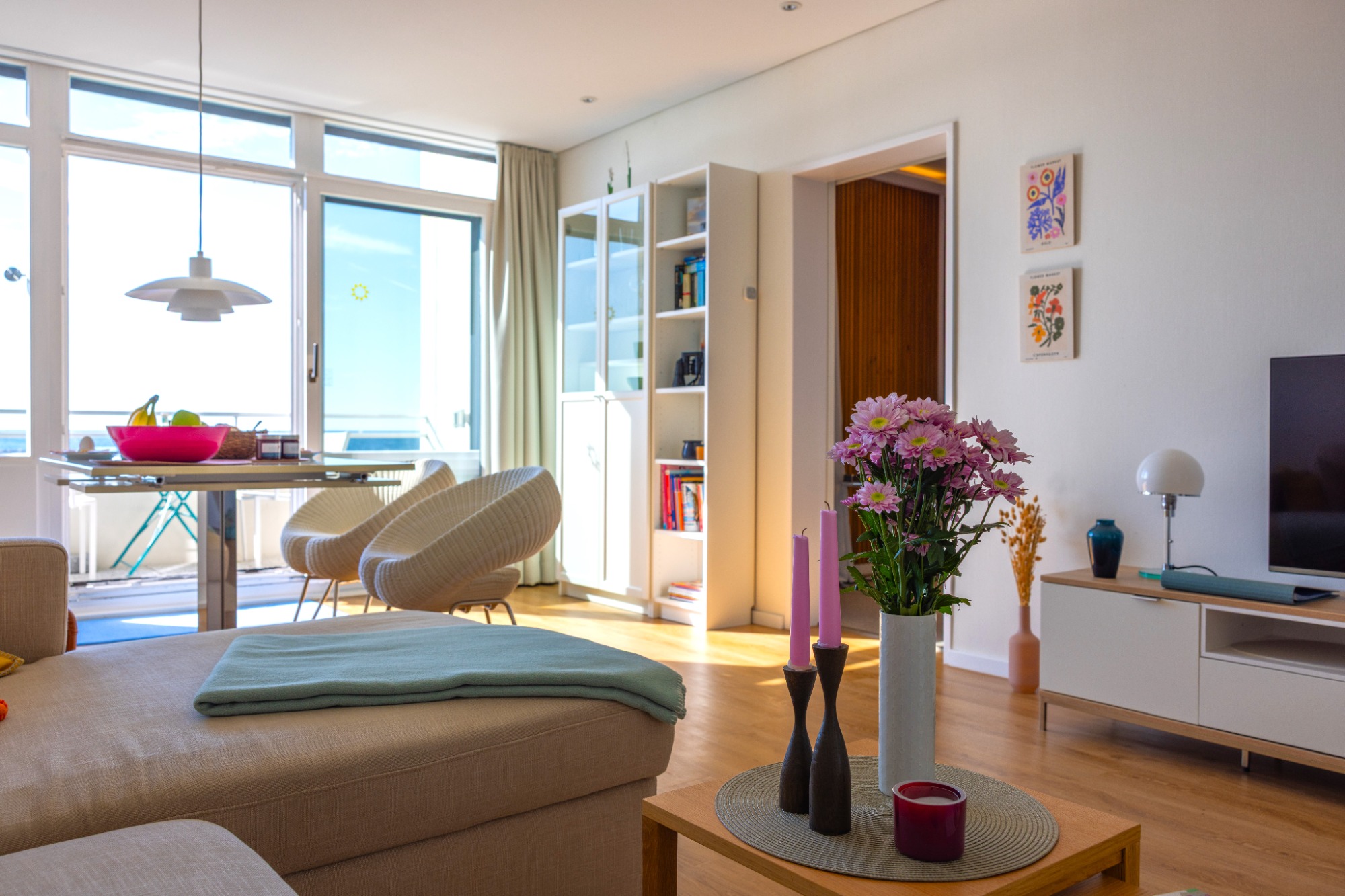 Entspannt-modernes Strandaparment Heller Wohnraum mit Sofas, Tisch, Pflanzen und TV, große Fenster mit Blick nach draußen.